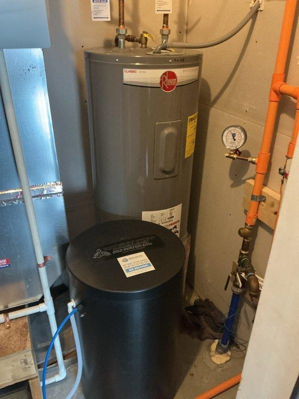 Responded to service call for leaking water heater in basement utility area. Upon inspection, found 50-gallon electric Rheem Classic series water heater with visible corrosion and active leak originating from bottom of tank. Unit was approximately 3 years old and showed signs of internal tank failure with rust-through condition. Documented existing installation including copper supply lines, brass fittings, flexible gas connections, and orange PEX distribution piping. Noted water softener system and Logic filtration equipment in immediate vicinity of failed heater. Assessed plumbing configuration including pressure gauge, wall-mounted connections, and hot water return line. Provided estimates for complete water heater replacement due to irreparable tank corrosion and leak severity.
