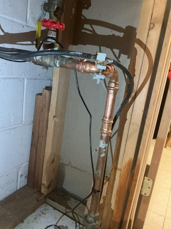 Responded to water leak emergency call for pinhole leak on copper supply line near water meter entry point in basement. Upon arrival, confirmed active leak on vertical copper pipe where main water line enters structure from ground. Leak was located on aged copper piping showing oxidation and patina, positioned adjacent to AO Smith ProLine water heater and water softening system. 

Performed repair of pinhole leak using appropriate copper pipe repair method. Applied white cloth wrapping at repair joints to secure connection points. Verified water meter shutoff valve was operational with red handle control. Tested repaired section under full water pressure to confirm leak was eliminated and no additional seepage was present at repair site or surrounding connections. Main water supply remained active throughout service as customer had not needed to shut off water to home prior to arrival.

Documented basement utility area configuration including proximity to water heater, filtration equip