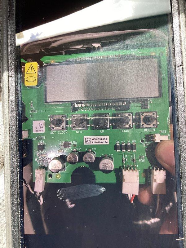 Completed warranty return trip to replace defective circuit board on water softener control head. Upon initial visit, identified faulty control board and ordered replacement part. Discovered incorrect part was received during this visit. Documented existing circuit board with LCD display, control buttons (SET CLOCK, NEXT, UP, DOWN, RE-GEN, TEST), and wire connections before removal. Removed defective board from metal housing and verified part specifications. Ordered correct replacement circuit board to complete repair on subsequent visit.