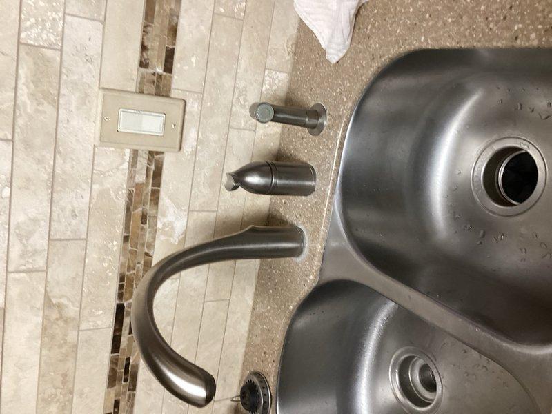 Responded to kitchen faucet leak complaint with water leaking onto countertop. Upon inspection, identified failed cartridge causing spout leak in existing bronze finish faucet assembly. Removed and replaced entire faucet system with new Moen three-piece fixture set including high-arc spout, side spray attachment, and soap dispenser component in coordinating oil-rubbed bronze finish to match existing décor. Installed on double-basin stainless steel sink with granite countertop. Tested all components for proper operation and verified leak resolution with no water discharge onto counter surface.