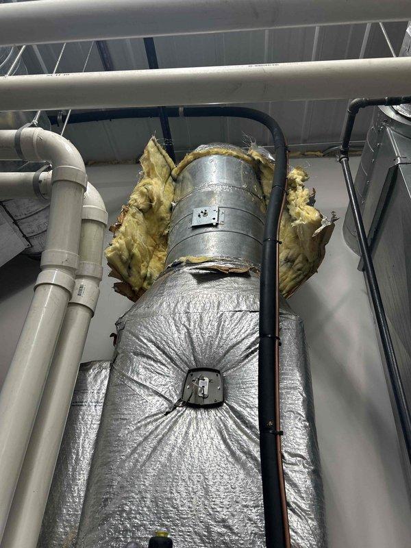 Responded to scheduled service call for damper assembly replacement in commercial HVAC system. Upon arrival, attempted to locate and replace two damper assemblies: one 12" x 18" side mount unit and one 14" round damper assembly. Unable to locate bathroom damper assembly during inspection of ductwork and mechanical spaces. Encountered significant difficulty accessing and removing the existing 14" round damper due to installation constraints and surrounding ductwork configuration. Photographed existing conditions showing large insulated cylindrical ductwork with reflective silver wrapping, yellow fiberglass insulation in deteriorated condition, white PVC piping, and black flexible conduit in confined ceiling space. Due to access challenges with the round damper removal and inability to locate the second damper, coordinated with dispatch to reschedule service call with original estimator to review installation plan and complete repairs.