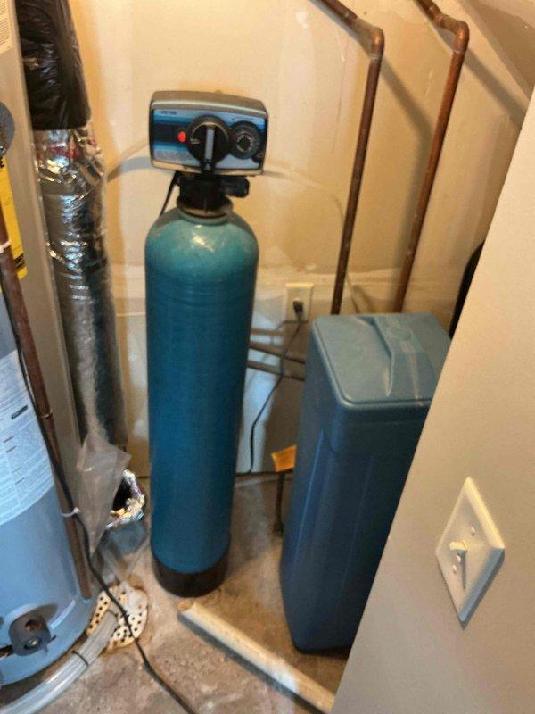 Responded to a water softener leak complaint at a townhome with an approximately 20-year-old water treatment system. Upon inspection, identified a slight leak on the existing teal-colored cylindrical water softener unit with digital control valve head. Assessed the condition of the aging system, including the pressure tank and associated copper piping connections. Documented the equipment configuration and leak source through photographic documentation for maintenance records and future service recommendations.