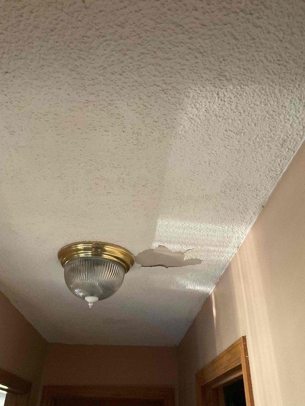 Responded to reported ceiling damage with peeling popcorn texture adjacent to ceiling-mounted light fixture outside frequently used bathroom. Conducted thorough inspection and testing to identify potential water leak source. Visual examination revealed compromised ceiling material with texture separation and hole formation near fixture mounting point, though no active water leak was detected at time of service. Documented ceiling damage conditions with photographs for record purposes.