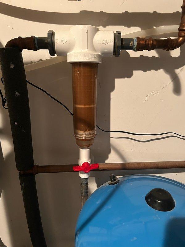 Serviced well water filtration system at pressure tank location in basement. Inspected sediment filter housing which showed significant discoloration and buildup due to extended service interval. Flushed filter housing, replaced dirty cartridge with new filtration media, and verified proper system operation. Provided estimate for installation of main water supply ball valve to improve system serviceability and allow for easier future maintenance shutdowns.