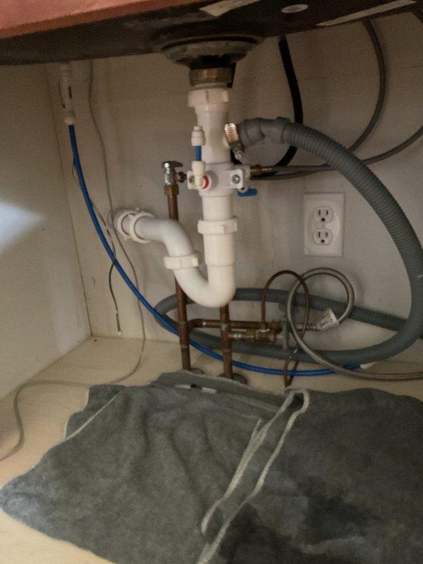 Responded to callback for persistent leak under kitchen sink following previous mitigation attempt. Upon arrival, inspected under-sink plumbing assembly and identified source of leak at strainer basket connection. Documented conditions with photographs showing white PVC P-trap configuration, copper water supply lines with shut-off valves, blue flexible supply hoses, and gray corrugated disposal/dishwasher drain line. Strainer basket seal confirmed as failure point causing continued water intrusion. Provided replacement quote to customer for strainer basket assembly to permanently resolve leaking issue.