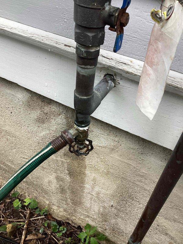 Responded to exterior hose bib leaking issue at residential property. Upon inspection, found outdoor spigot with deteriorated connections and corroded fittings causing water spray during operation. Identified that attached garden hose was contributing to leak at connection point. Removed garden hose from hose bib to isolate leak source. Assessed vertical pipe assembly and valve configuration, noting weathered components and evidence of previous repair attempts. Documented existing plumbing setup including multiple threaded connections, valve with control handles, and overall condition of exterior water supply system for recommended replacement or further repair.