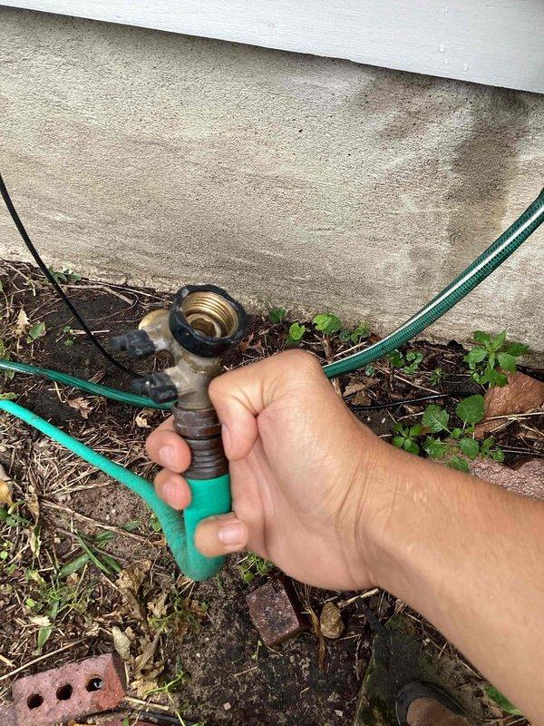 Responded to exterior hose bib leaking issue at residential property. Upon inspection, found outdoor spigot with deteriorated connections and corroded fittings causing water spray during operation. Identified that attached garden hose was contributing to leak at connection point. Removed garden hose from hose bib to isolate leak source. Assessed vertical pipe assembly and valve configuration, noting weathered components and evidence of previous repair attempts. Documented existing plumbing setup including multiple threaded connections, valve with control handles, and overall condition of exterior water supply system for recommended replacement or further repair.