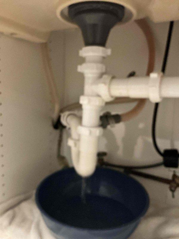 Responded to water leak under kitchen sink occurring intermittently without sink or dishwasher use. Upon inspection, identified backed-up drain line causing water to overflow from P-trap assembly when water softener discharged. Confirmed drain blockage was preventing proper drainage of softener discharge water, resulting in backup through sink drain system. Documented plumbing configuration including white PVC P-trap assembly, supply lines, and connections. Scheduled drain technician for follow-up service to clear obstruction and restore proper drainage function.