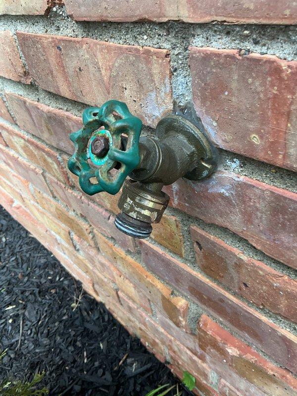 Responded to exterior water leak at outdoor hose bib mounted on brick wall. Upon arrival, found garden hose fused to the spigot's vacuum breaker assembly, preventing proper removal and causing significant water leakage. Water supply had been shut off prior to arrival. Successfully removed the compromised hose from the threaded connection and vacuum breaker. Inspected the brass spigot assembly with wheel-style valve handle for additional damage and assessed the condition of the fixture, noting weathering and oxidation consistent with age and outdoor exposure. Provided replacement options and cost estimates for the vacuum breaker assembly and potential full spigot replacement to prevent future leakage issues.