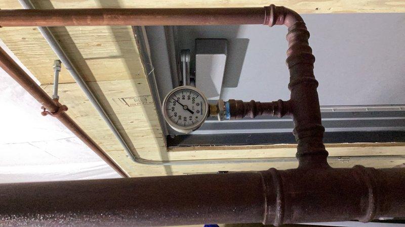 Completed comprehensive plumbing inspection of commercial facility including water heater relocation, PRV pressure verification, vacuum breaker testing, mixing valve functionality checks, water heater flushing and maintenance, faucet temperature and component testing, and eyewash station evaluation. Identified multiple deficiencies requiring attention: mixing valve in unisex bathroom requires adjustment, mixing valve and foot pedal at main handwashing sink need replacement, dishwashing room handwashing sink drain experiencing backup issues, and cold cartridge in lab handwashing sink requires replacement. Documented system conditions with photographs showing aged copper piping with heavy oxidation and pressure gauge monitoring, as well as portable handwashing station setup. Estimate to be prepared for identified repairs and component replacements.