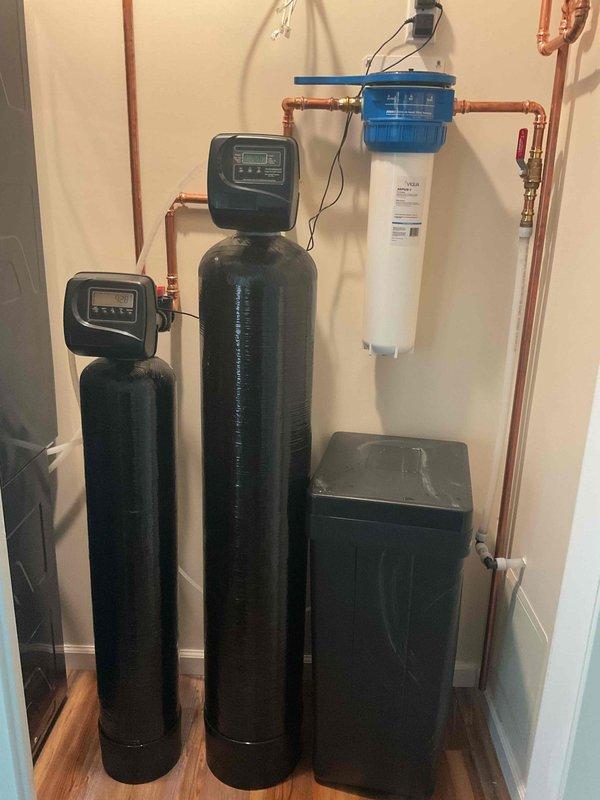 Completed manifold build for residential water treatment system consisting of three black cylindrical tanks with digital control heads. Installed isolation valves on copper piping to service tankless water heater. System includes two tall vertical softener/filtration units and one shorter rectangular tank, all connected via copper manifold with white filter housing featuring blue cap. Plumbing configuration incorporates multiple shut-off valves with red handles for individual component isolation and maintenance access. All connections secured and tested for proper operation.