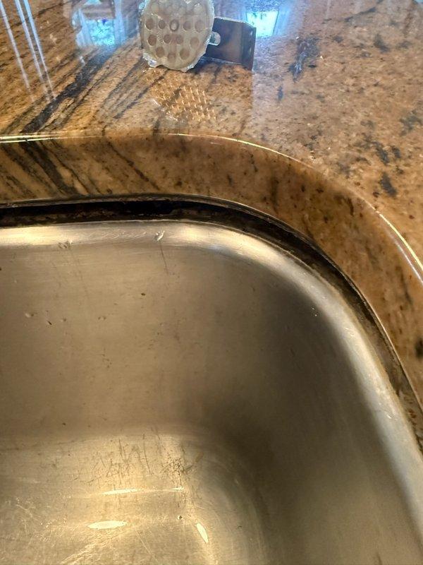 Responded to undermount kitchen sink that had dropped approximately 1/2 inch on one side following recent faucet replacement. Upon inspection, determined that adhesive holding sink mounting clamps had failed, causing clamps to detach from underside of granite countertop. Fabricated temporary support using cut pipe section to stabilize sink basin and prevent further displacement until permanent countertop repair can be completed by stone fabrication specialist.