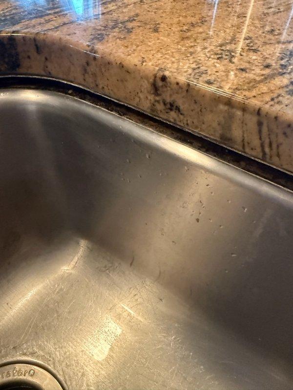 Responded to undermount kitchen sink that had dropped approximately 1/2 inch on one side following recent faucet replacement. Upon inspection, determined that adhesive holding sink mounting clamps had failed, causing clamps to detach from underside of granite countertop. Fabricated temporary support using cut pipe section to stabilize sink basin and prevent further displacement until permanent countertop repair can be completed by stone fabrication specialist.