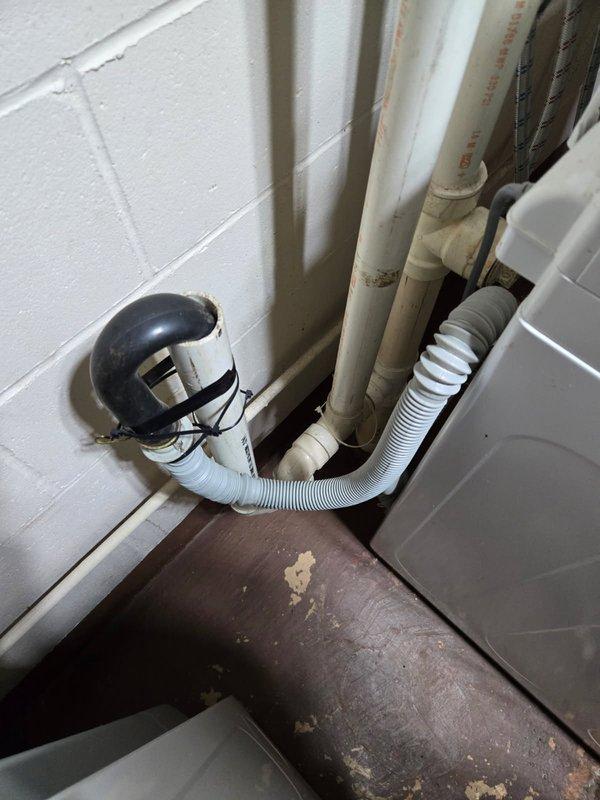 Responded to floor drain backup occurring during laundry operation. Attempted to clear blockage by snaking both the floor drain and the main stack line. Inspected laundry drain plumbing system including P-trap connections and drainage assembly beneath fixtures. Work performed in utility area with exposed plumbing infrastructure along cinder block walls.