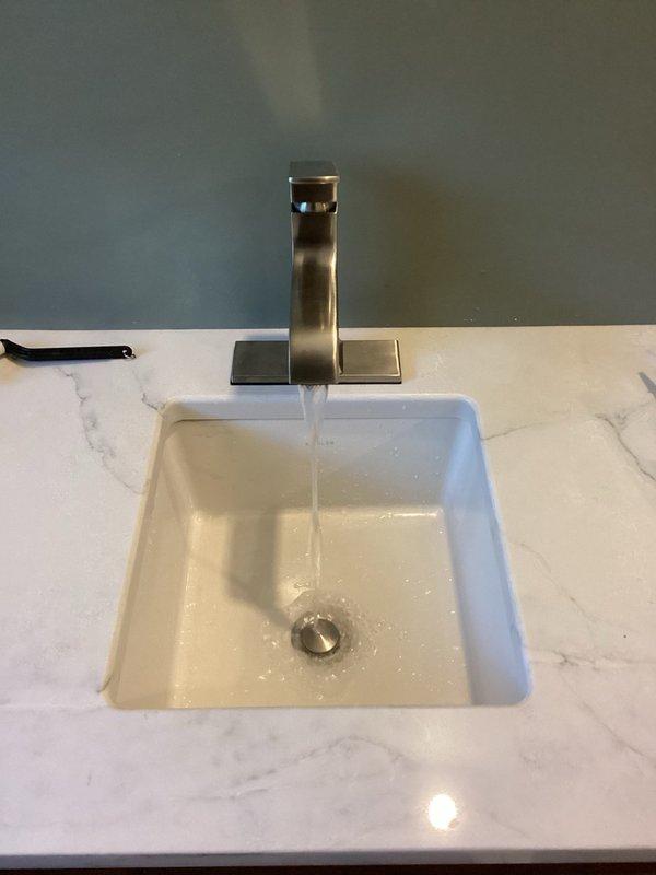 Successfully installed three customer-supplied bathroom fixtures including two lavatory faucets and one toilet. The first installation featured a contemporary brushed nickel faucet with angular geometric design mounted to a white countertop with gray marble veining, paired with a rectangular undermount sink. The second installation consisted of a three-piece widespread faucet in brushed finish with separate handle controls and curved spout, installed with an oval undermount lavatory. All fixtures were tested thoroughly for proper operation and leak-free performance by filling and draining each sink multiple times, running continuous water flow for ten minutes, and flushing the toilet ten times. No leaks were detected at any connections. All fixtures left in full working order.