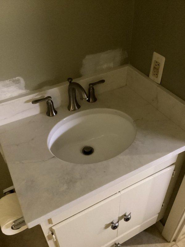 Successfully installed three customer-supplied bathroom fixtures including two lavatory faucets and one toilet. The first installation featured a contemporary brushed nickel faucet with angular geometric design mounted to a white countertop with gray marble veining, paired with a rectangular undermount sink. The second installation consisted of a three-piece widespread faucet in brushed finish with separate handle controls and curved spout, installed with an oval undermount lavatory. All fixtures were tested thoroughly for proper operation and leak-free performance by filling and draining each sink multiple times, running continuous water flow for ten minutes, and flushing the toilet ten times. No leaks were detected at any connections. All fixtures left in full working order.