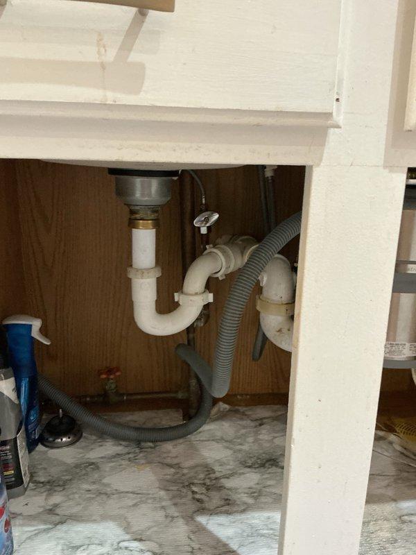 Responded to clogged kitchen sink complaint. Upon inspection, identified blockage caused by accumulation of grease and egg shells in the drain line. Accessed plumbing system beneath sink, which included chrome drain basket, white PVC P-trap assembly, and garbage disposal unit with associated drainage components. Utilized rigid cable machine to clear obstruction from drain line, successfully restoring proper flow and drainage function. Documented existing cabinet conditions including water staining on cabinet floor and plumbing configuration. All drain components tested and verified operational upon completion of cleaning procedure.
