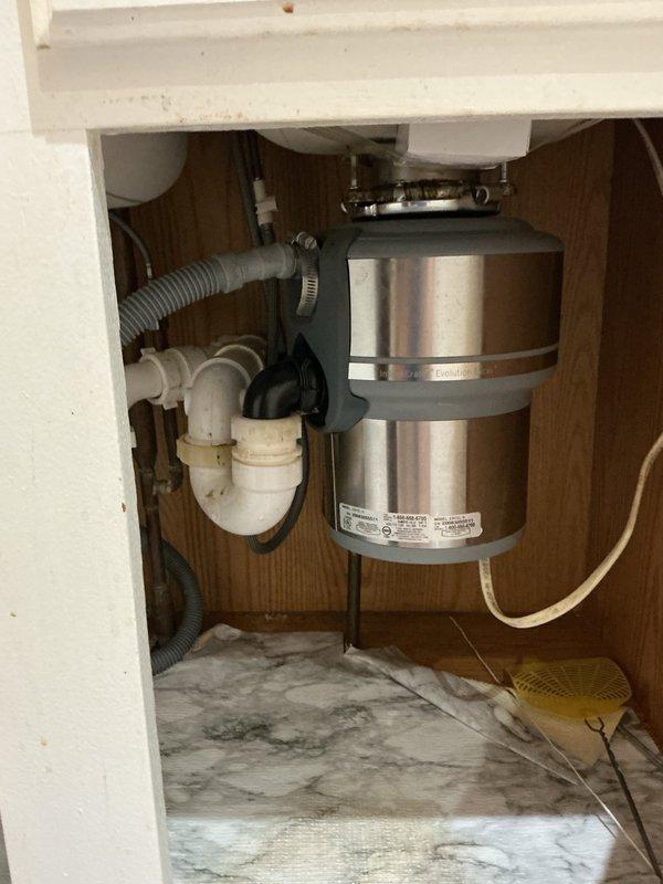 Responded to clogged kitchen sink complaint. Upon inspection, identified blockage caused by accumulation of grease and egg shells in the drain line. Accessed plumbing system beneath sink, which included chrome drain basket, white PVC P-trap assembly, and garbage disposal unit with associated drainage components. Utilized rigid cable machine to clear obstruction from drain line, successfully restoring proper flow and drainage function. Documented existing cabinet conditions including water staining on cabinet floor and plumbing configuration. All drain components tested and verified operational upon completion of cleaning procedure.