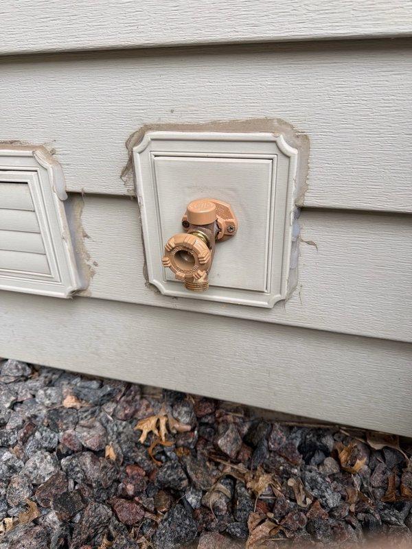 Replaced exterior hose bib with new fixture mounted on white panel against building siding, utilizing copper connections. Installed access panel in basement floor opening to provide future serviceability to the water supply line, which includes PVC piping with shut-off valve and proper insulation at the floor level.