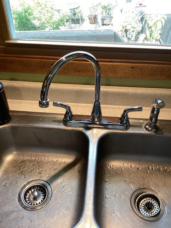 Installed customer-supplied chrome kitchen faucet with dual handles and side sprayer attachment on double-basin stainless steel sink. Verified proper water flow, tested for leaks, and ensured all connections were secure beneath the sink cabinet.