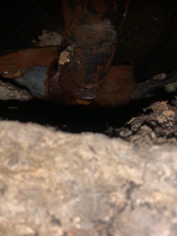 Responded to service call for boiler system with frozen and damaged hydronic heating piping. Upon inspection, found severely corroded 1" black iron pipe with perforation and breach in inaccessible location serving baseboard heating loop in rear section of main level. Corrosion and freeze damage resulted from loop being shut off during previous winter season. Provided estimate for repair solution involving isolation of damaged section by installing shut-off ball valves at accessible basement location, abandoning deteriorated black iron piping in place, and running new PEX tubing to supply side of existing baseboard units. Main boiler system confirmed operational and serving remaining radiators throughout property. Documented existing conditions with photographs showing extent of pipe corrosion, perforation damage, and confined crawl space access constraints.