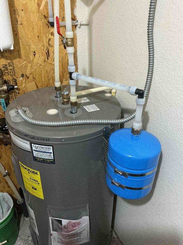 Installed a whole home carbon filtration system with blue housing and white PVC piping connections mounted to the utility room wall. Added a blue expansion tank to the existing Gunderson water heater, securing it with metal clamps and integrating it into the plumbing system with proper brass fittings, valves, and flexible connectors to protect against thermal expansion and ensure code compliance.