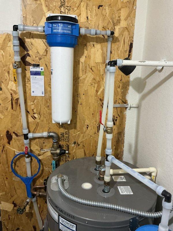 Installed a whole home carbon filtration system with blue housing and white PVC piping connections mounted to the utility room wall. Added a blue expansion tank to the existing Gunderson water heater, securing it with metal clamps and integrating it into the plumbing system with proper brass fittings, valves, and flexible connectors to protect against thermal expansion and ensure code compliance.