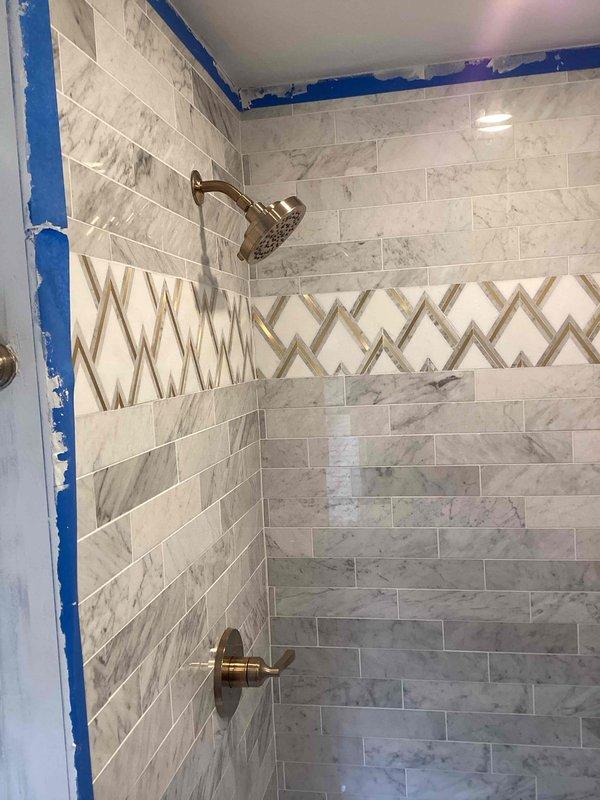 Completed trim-out of shower valves in newly tiled shower enclosure featuring gray marble-pattern tiles with decorative chevron accent band. Installed brushed bronze shower head and single-handle control valve, coordinating with tile design. Additionally replaced two lavatory faucets with new units including pop-up drain assemblies per owner's verbal authorization.