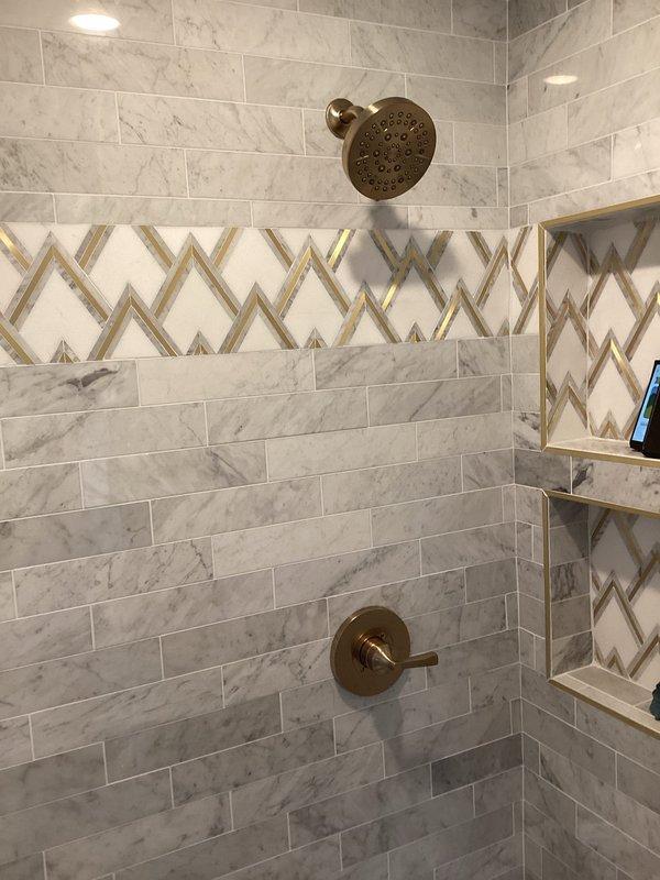 Completed trim-out of shower valves in newly tiled shower enclosure featuring gray marble-pattern tiles with decorative chevron accent band. Installed brushed bronze shower head and single-handle control valve, coordinating with tile design. Additionally replaced two lavatory faucets with new units including pop-up drain assemblies per owner's verbal authorization.
