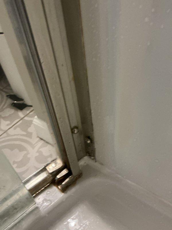 Responded to shower leak complaint and performed on-site inspection of bathroom enclosure. Upon arrival, identified the source of water leakage as a deteriorated shower door assembly with compromised metal hardware components. The glass/acrylic door panel exhibited significant rust and corrosion on the bottom track, latch mechanism, and mounting brackets, which had caused seal failure and allowed water to escape during shower use.

Documented the extensive oxidation damage on the metallic door frame components, including the rectangular catch mechanism and base track system. The rust staining extended onto the white tiled surfaces, indicating prolonged moisture exposure and hardware deterioration. Assessed the condition of the door guide mechanisms and floor junction where the enclosure meets the decorative tile flooring. The corroded metal components had compromised the door's ability to maintain a watertight seal, resulting in active water leakage during operation.