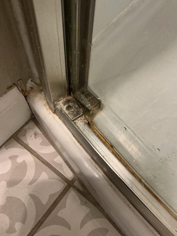 Responded to shower leak complaint and performed on-site inspection of bathroom enclosure. Upon arrival, identified the source of water leakage as a deteriorated shower door assembly with compromised metal hardware components. The glass/acrylic door panel exhibited significant rust and corrosion on the bottom track, latch mechanism, and mounting brackets, which had caused seal failure and allowed water to escape during shower use.

Documented the extensive oxidation damage on the metallic door frame components, including the rectangular catch mechanism and base track system. The rust staining extended onto the white tiled surfaces, indicating prolonged moisture exposure and hardware deterioration. Assessed the condition of the door guide mechanisms and floor junction where the enclosure meets the decorative tile flooring. The corroded metal components had compromised the door's ability to maintain a watertight seal, resulting in active water leakage during operation.