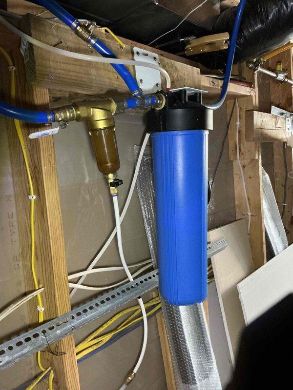 Completed comprehensive plumbing inspection of older farmhouse with extensive DIY installations. Documented existing water filtration system in crawl space, including blue cylindrical filter housing with brass fittings, sediment filter with clear bowl, and PEX piping distribution throughout structure. All accessible plumbing components, connections, and routing were visually examined and photographed for maintenance records.