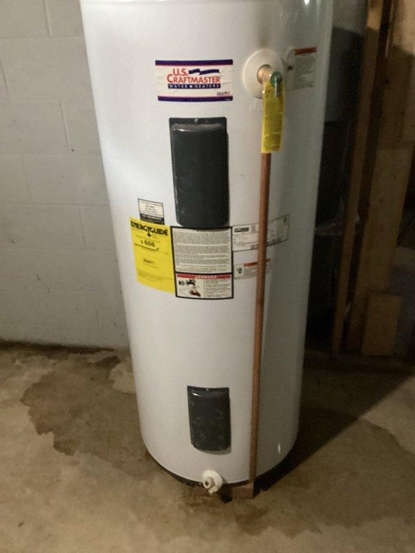 Responded to service call for leaking water heater with visible water accumulation on floor. Upon inspection, identified an 80-gallon U.S. Craftmaster electric water heater actively leaking from the bottom of the tank, indicating internal tank failure. Unit was still producing hot water at time of assessment. Documented existing conditions with photographs showing the water heater installation in basement utility area, copper supply lines, yellow-handled shut-off valve, and water staining on concrete floor surrounding the base of the unit. Provided customer with detailed estimate for complete water heater replacement due to non-repairable tank failure.