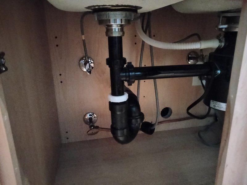 Responded to callback regarding kitchen and bathroom shutoff valve replacement following previous leak repair service. Inspected kitchen sink cabinet plumbing assembly, including copper supply lines, chrome shutoff valves, black PVC P-trap configuration, and stainless steel drain basket strainer. Documented existing valve conditions under kitchen sink with two chrome shutoff valves mounted on back panel with black flexible supply lines. Also assessed bathroom shutoff valve requiring replacement. Confirmed availability of necessary valve replacement parts and provided estimates for shutoff valve replacement work at both locations for customer consideration.