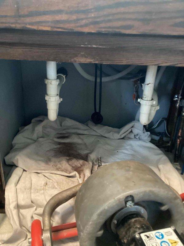 Responded to a recurring kitchen sink drain blockage that had been previously serviced less than a month prior. Accessed the drain system through the cabinet base and utilized mechanical cable equipment to clear the obstruction in the drain line. Upon completion, tested water flow through both basins of the double-bowl stainless steel sink to verify proper drainage and confirm the blockage was fully removed.
