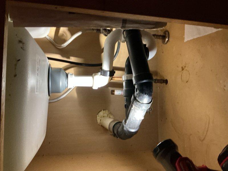 Responded to service call for leak under kitchen sink. Upon inspection, identified leak in drain assembly beneath sink with garbage disposal. Examined P-trap connections and tailpiece assembly for source of water intrusion. Assessed condition of existing drain piping system, noting mixed materials including PVC and corrugated flexible tubing. Provided estimate for complete replacement of kitchen drain piping system to resolve leak issue and upgrade to code-compliant materials. Documented existing plumbing configuration with photographs showing garbage disposal connection, P-trap assembly, and cabinet condition for repair proposal.