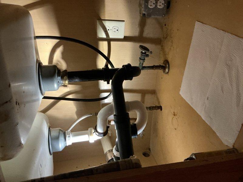 Responded to service call for leak under kitchen sink. Upon inspection, identified leak in drain assembly beneath sink with garbage disposal. Examined P-trap connections and tailpiece assembly for source of water intrusion. Assessed condition of existing drain piping system, noting mixed materials including PVC and corrugated flexible tubing. Provided estimate for complete replacement of kitchen drain piping system to resolve leak issue and upgrade to code-compliant materials. Documented existing plumbing configuration with photographs showing garbage disposal connection, P-trap assembly, and cabinet condition for repair proposal.