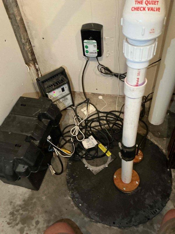 Performed comprehensive inspection of residential sump pump system per customer request. Examined submersible pump unit installed in concrete pit, verified proper operation and positioning, and assessed water level conditions. Inspected complete discharge piping system including vertical PVC discharge line with quiet check valve, confirmed secure mounting and proper connections at basin interface. Evaluated battery backup system components including wall-mounted control unit, battery charger, and backup pump assembly to ensure emergency operation capability. All electrical connections, power cords, and control systems were visually inspected for proper installation and functionality. System found to be operational with all components functioning as designed.