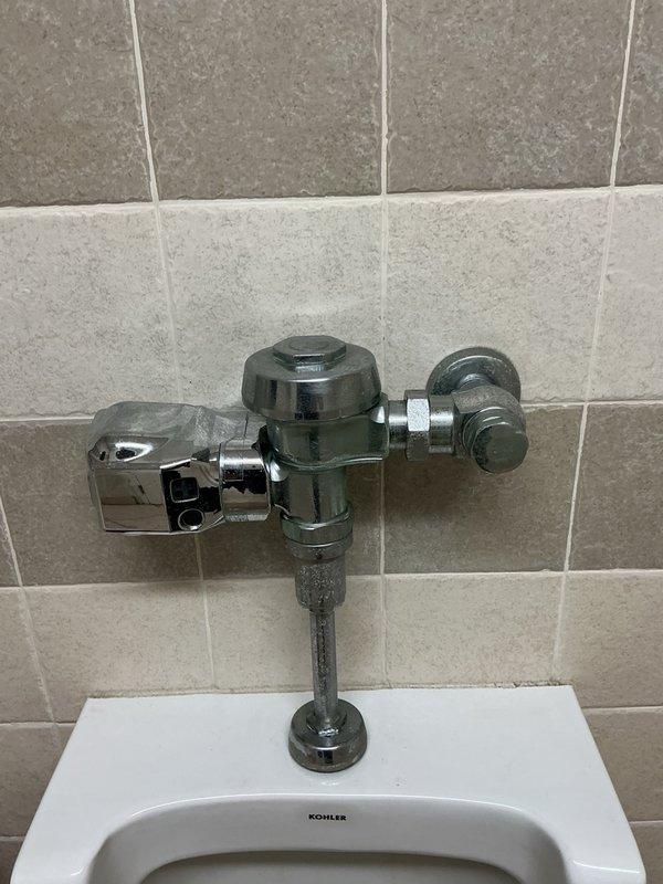 Responded to commercial urinal continuously running and flushing on its own. Upon inspection, identified dirty sensor on automatic flush valve assembly causing false activation. Cleaned sensor on Kohler wall-mounted urinal flush valve system, restoring proper operation and resolving continuous water flow issue.