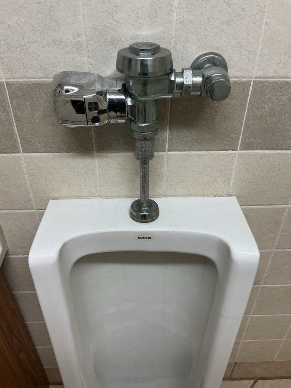 Responded to commercial urinal continuously running and flushing on its own. Upon inspection, identified dirty sensor on automatic flush valve assembly causing false activation. Cleaned sensor on Kohler wall-mounted urinal flush valve system, restoring proper operation and resolving continuous water flow issue.