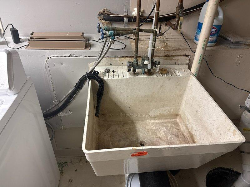 Responded to laundry tub drainage failure caused by cracked PVC drain tee fitting on the discharge line. Upon inspection, identified multiple deteriorated PVC fittings in the drain assembly connecting to the main sewer line prior to its entry into the concrete foundation. The drain tee on the right side of the laundry tub exhibited structural failure, and additional compromised sections were found in the taped areas of the drainage system. Temporarily secured the damaged sections to prevent sewer gas infiltration and allow completion of the washer cycle. Scheduled complete replacement of the laundry tub assembly including basin, faucet, supply stops, and drain piping to restore proper functionality and eliminate further drainage issues.