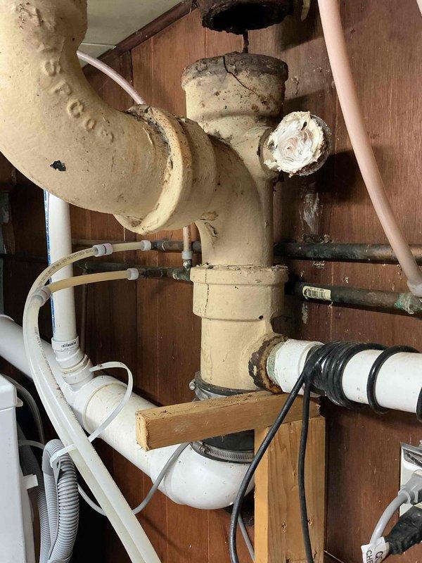 Responded to a drainage emergency in a 1941-era home where a deteriorated cast iron waste pipe had dropped approximately 5 inches from its original position, causing complete backup of all drains into the basement. Upon inspection, found the aged cast iron pipe had failed at a joint connection, with visible signs of corrosion and structural compromise. Documented the extent of damage through photographs showing the displaced pipe, surrounding water stains on wooden structural members, and temporary foam sealant material at the floor penetration point. The failed section exhibited significant discoloration and deterioration consistent with 80+ year-old cast iron plumbing. Assessed the existing plumbing configuration, which included a mix of original cast iron, PVC drain lines, and various connecting components mounted to wooden backing. Provided a comprehensive estimate for complete pipe repair and replacement of the compromised cast iron section to restore proper drainage function and 