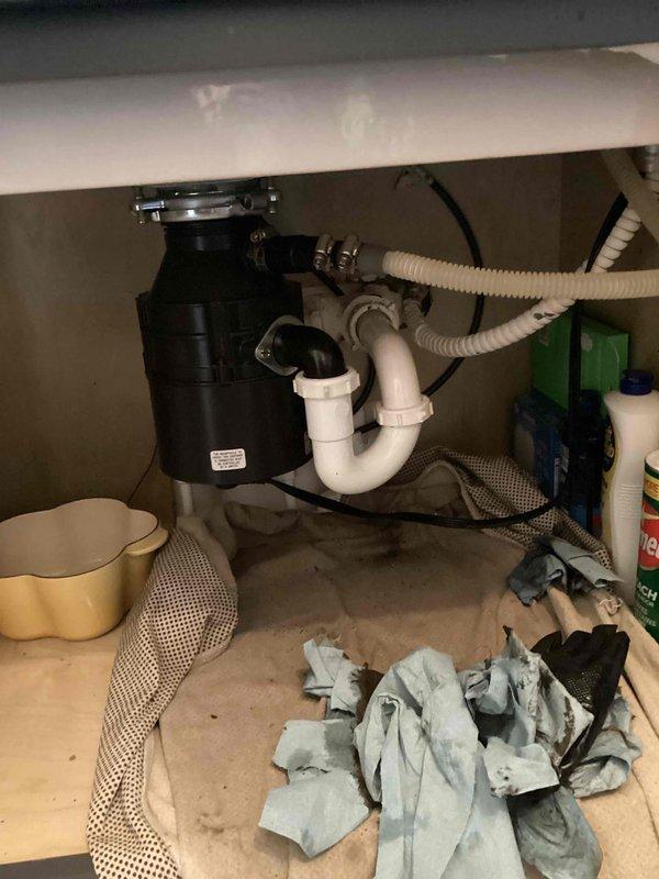 Responded to kitchen sink backup caused by clogged drain line. Upon arrival, found sink draining slowly with standing water and debris accumulation around drain opening. Customer had previously removed partial blockage from P-trap but drainage issue persisted. Inspected under-sink plumbing assembly including garbage disposal unit, PVC drain pipes, and P-trap configuration. Utilized drain cable to clear main drain line, successfully removing remaining obstruction that was causing backup. Verified proper drainage flow through sink and disposal unit. Tested garbage disposal operation to ensure functionality was restored. System now draining properly with no backup issues.