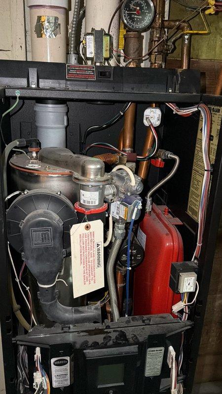 Responded to no-heat call on 3-year-old boiler system with radiator heat distribution. Upon inspection, found control board sustained water damage from overhead leak, resulting in complete loss of power to unit and non-functional display screen. This marked the second occurrence of water-related damage to the electrical components. Documented system condition showing internal mechanical compartment with expansion tank, circulator pump, copper piping, and compromised control board requiring replacement to restore heating operation.