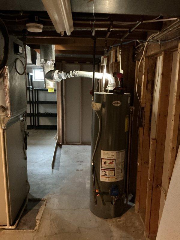 Performed inspection of 8-year-old tanked water heater in response to venting concerns. Upon examination, identified improper venting configuration with back-pitched vent pipe, which violates code requirements and creates potential for condensate accumulation and exhaust gas spillage. Documented existing conditions with photographs showing water heater installation alongside HVAC system in basement mechanical room. Verified water heater connections including gas supply lines with red valve handles and blue water connections at base of unit. Assessed overall system condition and surrounding mechanical equipment including furnace with white PVC intake/exhaust vents and metal ductwork. Provided estimates for new water heater replacement to address venting deficiencies and ensure proper code-compliant installation.