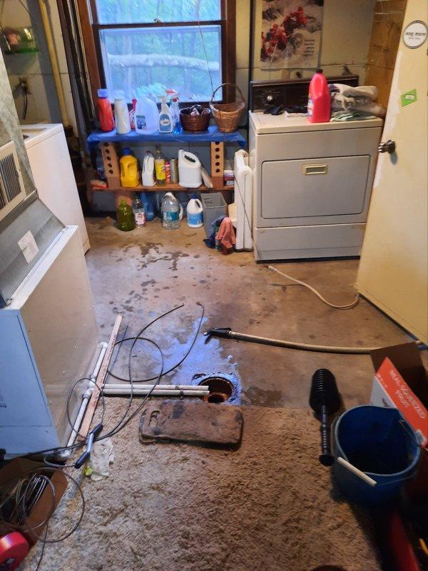 Responded to basement floor drain backup with water accumulation in laundry area. Located soft plug obstruction approximately 10 feet from access point in drainage line. Successfully cleared blockage using mechanical snake equipment, restoring proper drainage flow. Obstruction was positioned beyond 90-degree pipe bend, requiring specialized technique to navigate turn and reach clog. Water damage visible on laundry room floor from backup prior to service. Drain tested and confirmed flowing freely after clearance completed.