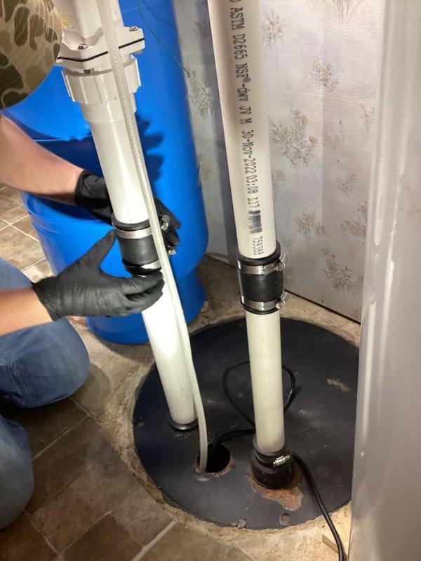 Responded to service call for non-operational sump pump and sewage ejector system. Upon inspection, determined that the sewage ejector pump had failed despite being a relatively recent installation. Replaced the defective sewage ejector pump under manufacturer warranty. Installation included proper mounting of two white PVC discharge pipes with rubber couplings through the floor penetration, secured with a black rubber boot seal. Installed red shut-off valve and control box on pipe assembly for system operation and maintenance access. Tested system functionality and verified proper pump operation and discharge flow.