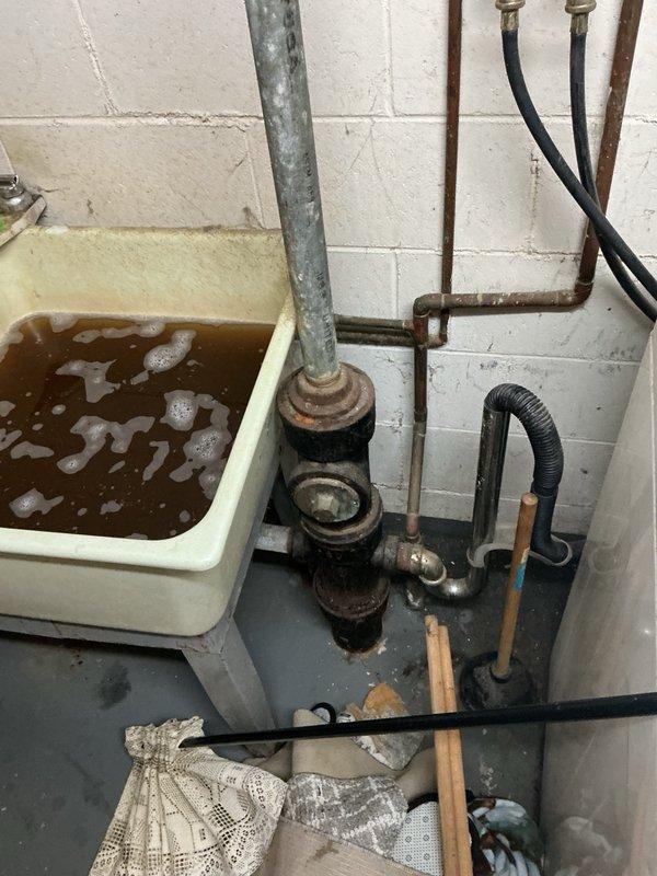 Responded to call for backing up basement and kitchen sinks in 1966 residential property with galvanized plumbing. Upon arrival, found kitchen sink drain severely caked with grease buildup and multiple leaking compression nuts under sink. Basement utility sink contained standing water with visible backup. Observed heavily corroded and rusted galvanized piping throughout drain system with advanced deterioration at joints and fittings. Attempted mechanical snaking to clear blockage but was unsuccessful due to extent of buildup and pipe deterioration. Documented conditions with photos showing corroded plumbing assembly, standing water in utility sink, and compromised drain connections. Advised that existing galvanized drain system has reached end of service life and requires complete replacement to resolve drainage issues and prevent further leaks.