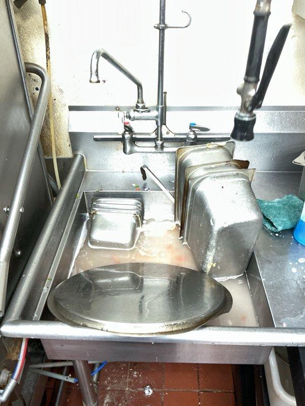 Responded to a recurring drain clog in the commercial kitchen where wastewater backed up through the floor drain during dishwasher and adjacent sink operation. Inspection confirmed standing, debris-laden water in the sink basin and a blockage in the main drain line below the floor, indicating a restriction between the dishwasher discharge and the grease trap. The affected drain line was assessed and documented, and a drain specialist will return to clear the main line obstruction and fully restore flow.