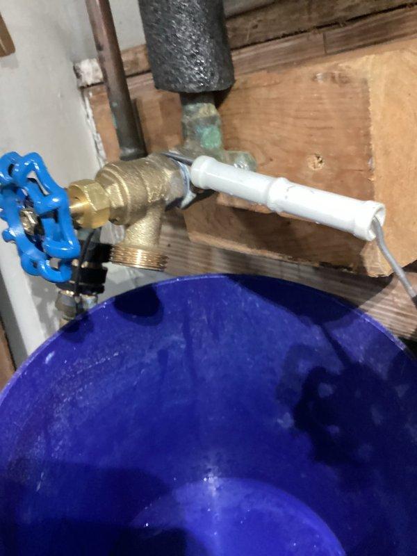Responded to callback for dripping valve in garage following recent water heater replacement. Upon arrival, inspected gate valve with blue handle connected to water supply system near pressure tank. Valve exhibited continuous dripping after being cycled open and closed. Assessed brass fittings, connections, and valve assembly for potential seat damage or debris causing improper seal. Documented plumbing configuration including pressure tank, gate valves, drain setup with hose connection to bucket, and associated piping. Photographed installation to record current condition and identify source of leak for repair recommendations.